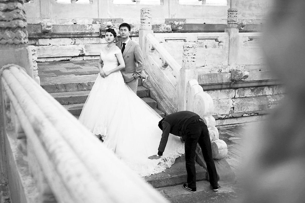 a couple taking wedding picture in front of the temple while a men is fixing the brides dress