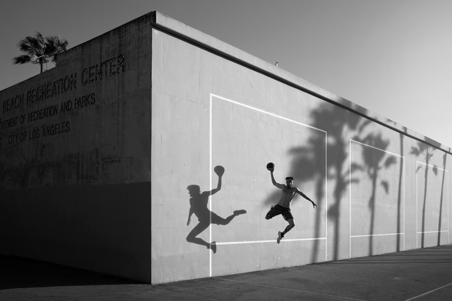 Guy jumping in front of the venice beach tennis court with his shaddow on the wall